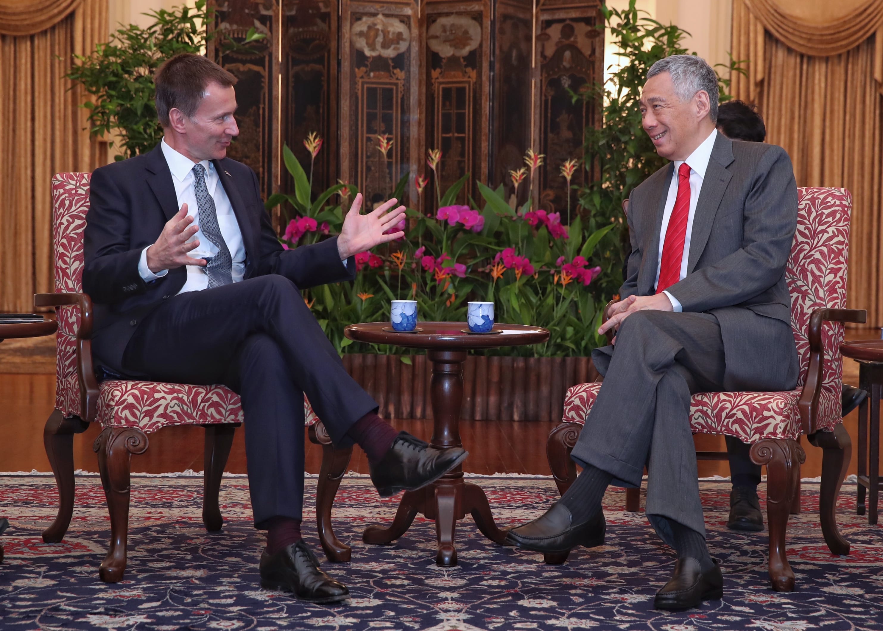 Two men in suits sit in ornate chairs, facing each other in a room decorated with plants and a painted screen.